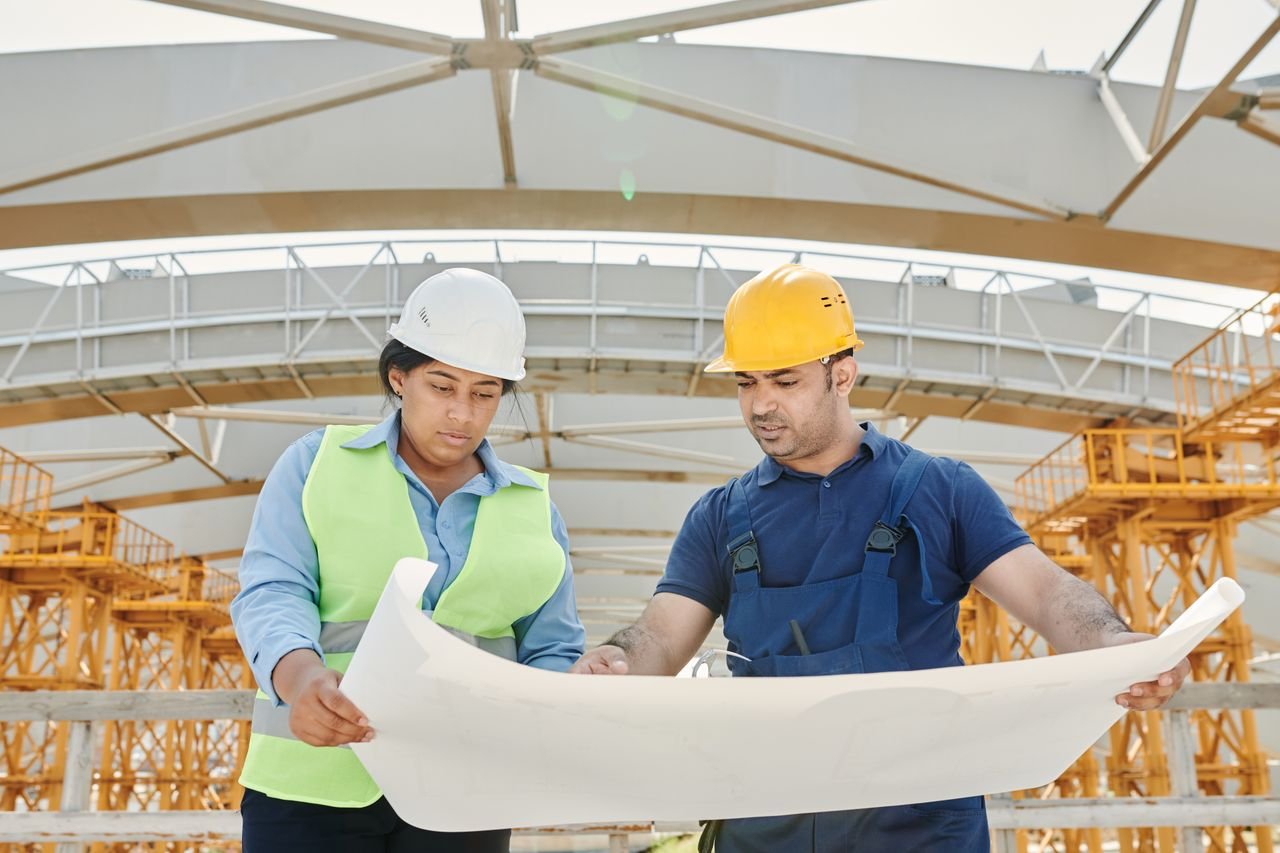 collection-01 Male and female engineers discussing blueprints at a construction site, promoting teamwork and planning.